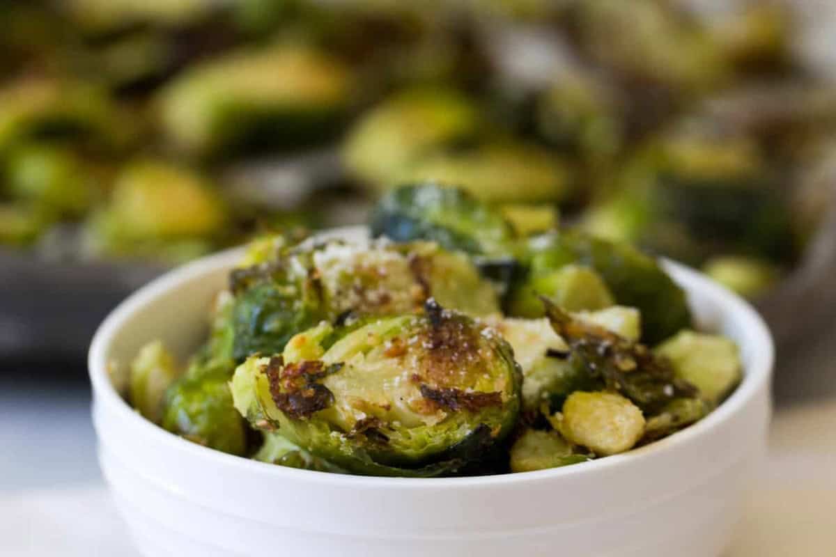 A white bowl filled with roasted Brussels sprouts topped with grated cheese, with a blurred background of more Brussels sprouts.