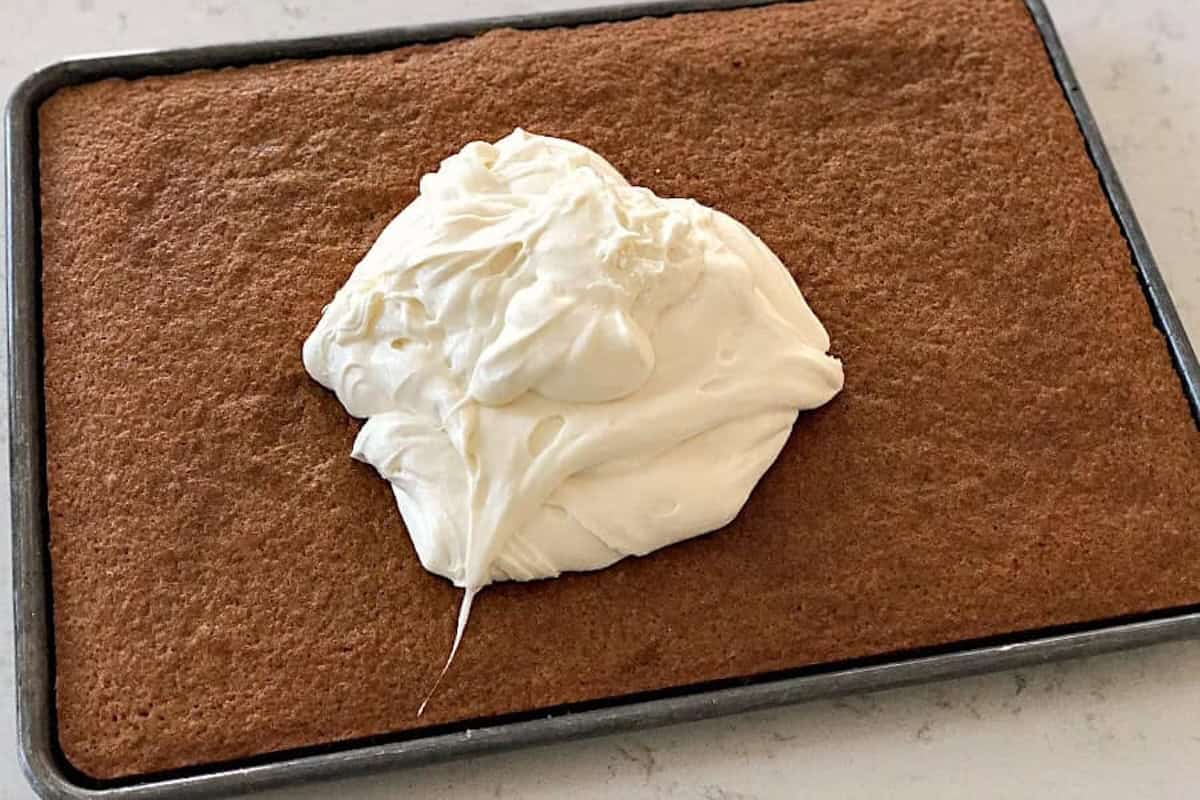 A rectangular cake in a baking pan with a dollop of white frosting on top, ready to be spread.