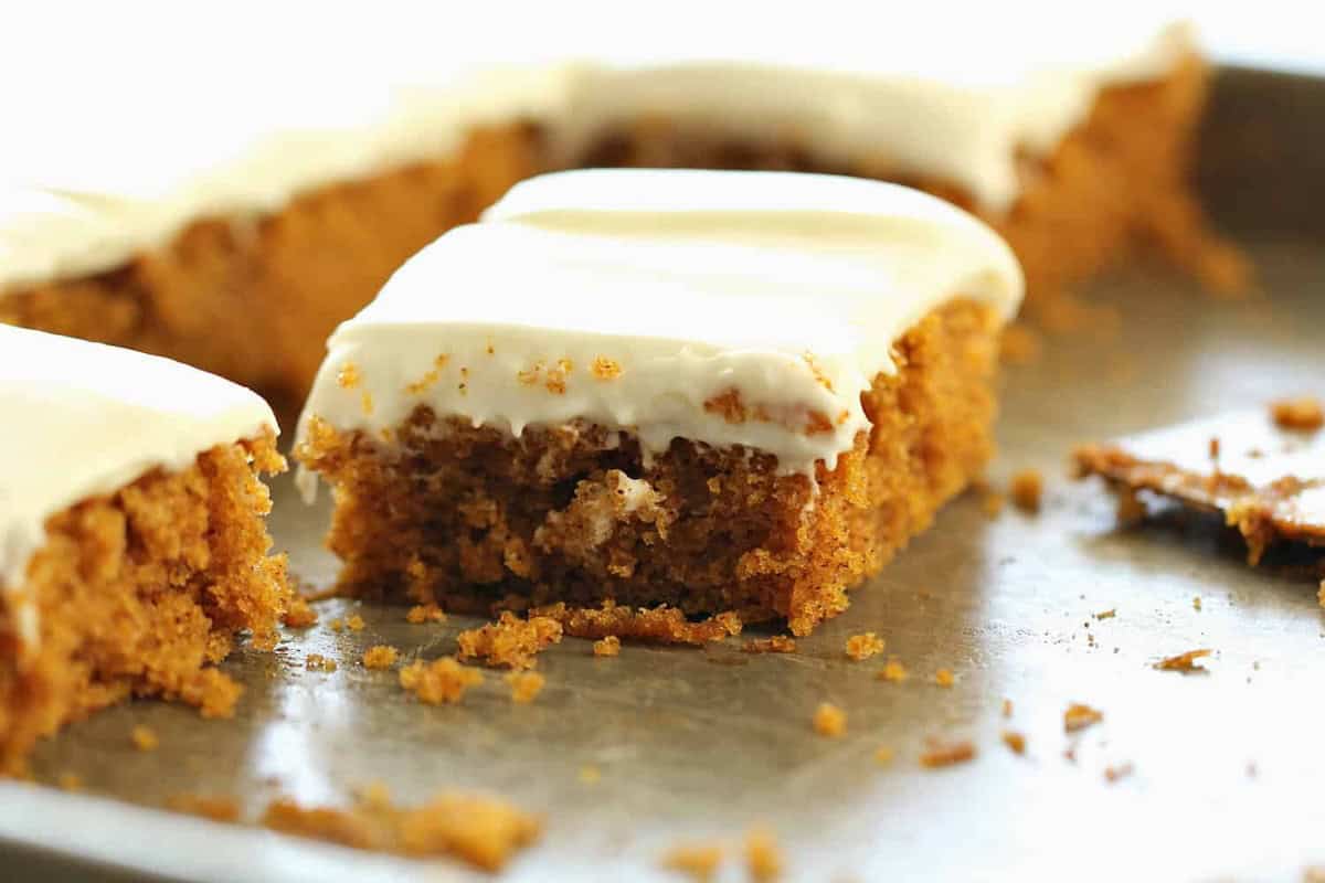 A close-up of a rectangular slice of carrot cake with cream cheese frosting on a baking sheet, with more slices in the background.