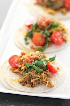 Close-up of three soft tacos on a plate filled with shredded meat, chopped tomatoes, leafy greens, and cheese.