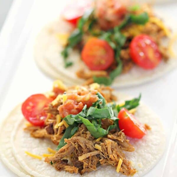 Close-up of three soft tacos on a plate filled with shredded meat, chopped tomatoes, leafy greens, and cheese.