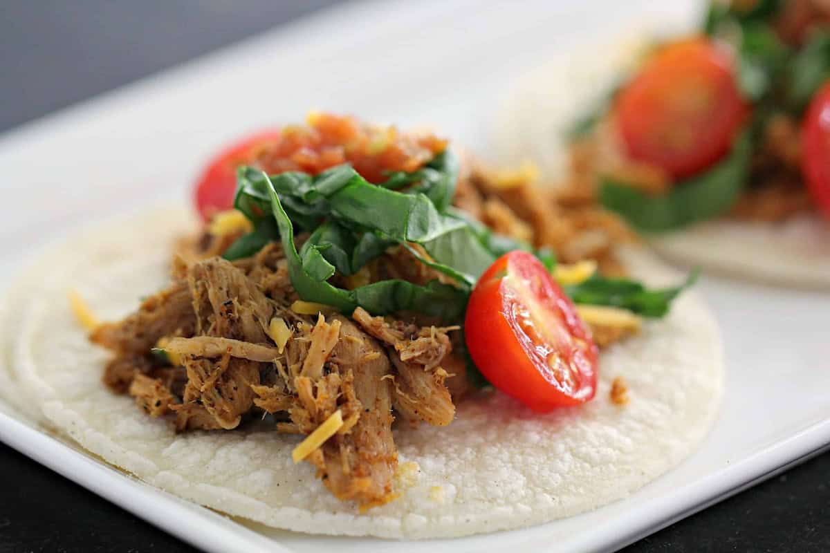 Close-up of two tacos on a white plate, each filled with shredded meat, chopped greens, and halved cherry tomatoes.