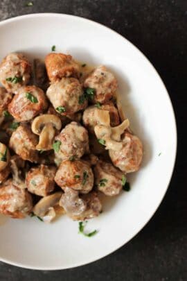 A white plate filled with meatballs and mushrooms in a creamy sauce, garnished with chopped parsley.