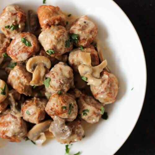 A white plate filled with meatballs and mushrooms in a creamy sauce, garnished with chopped parsley.