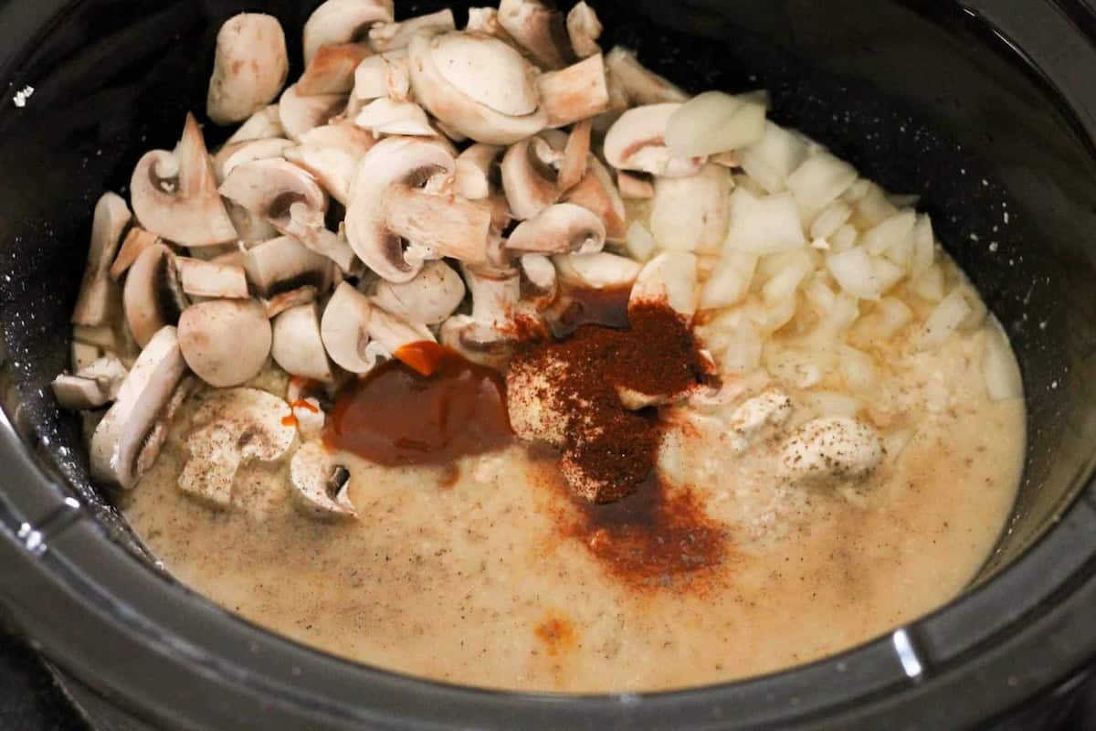 A slow cooker containing chopped mushrooms, diced onions, spices, and liquid mixture.