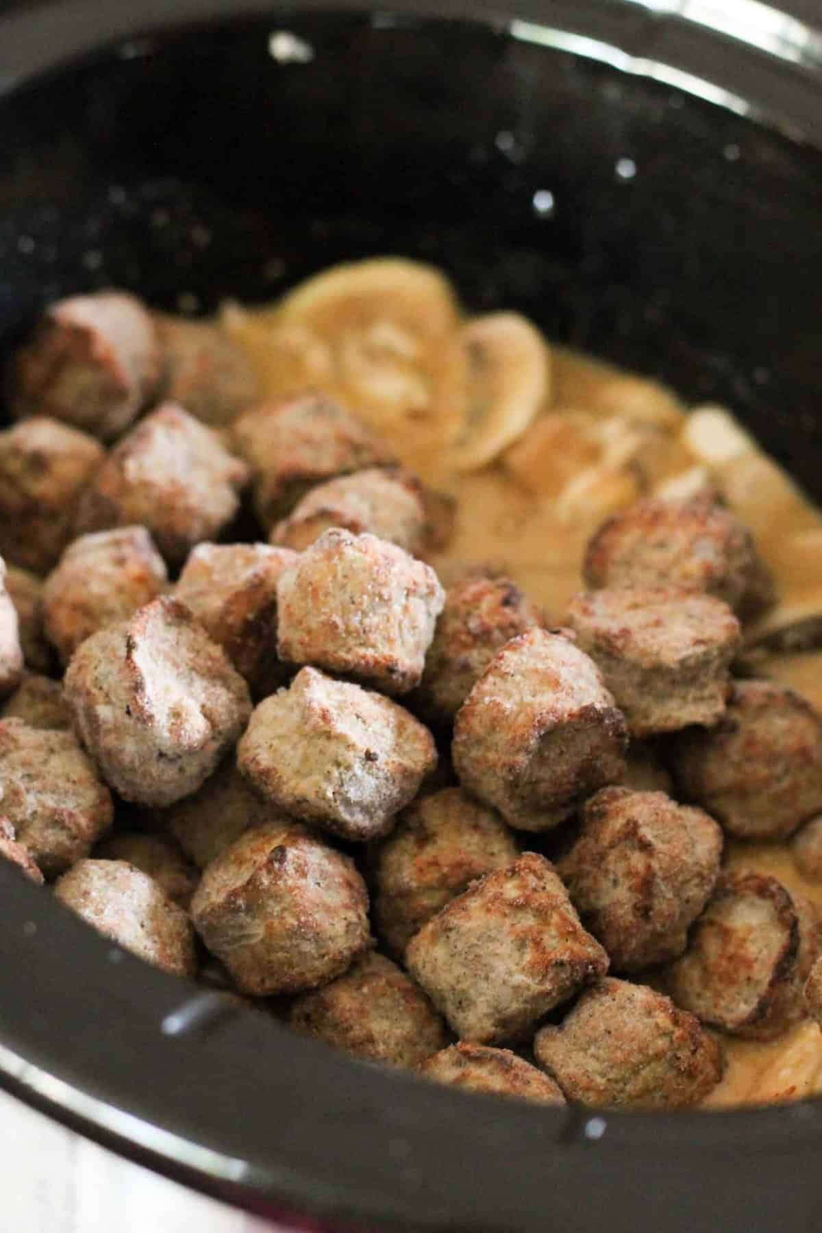 A slow cooker filled with meatballs in a creamy sauce with sliced mushrooms.