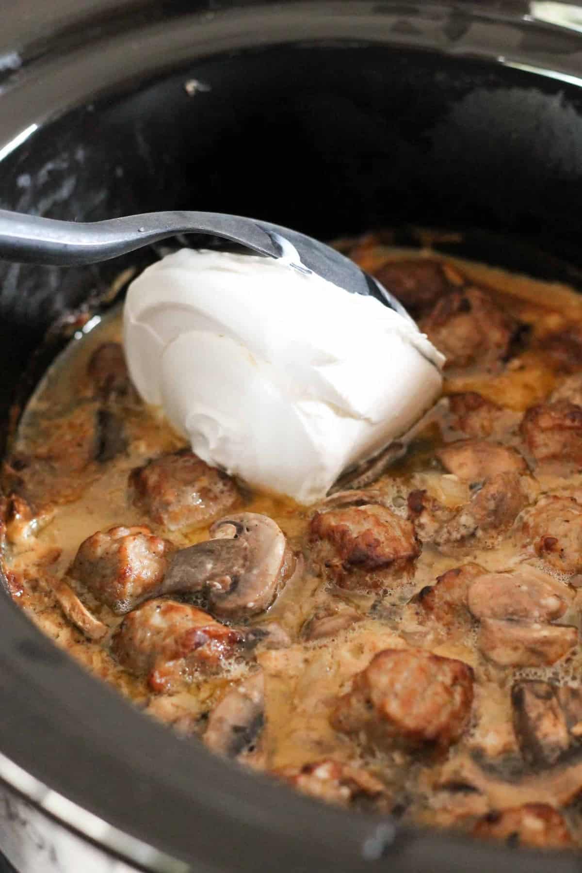 A spoonful of cream is being added to a slow cooker filled with sausage pieces and mushrooms in a creamy sauce.