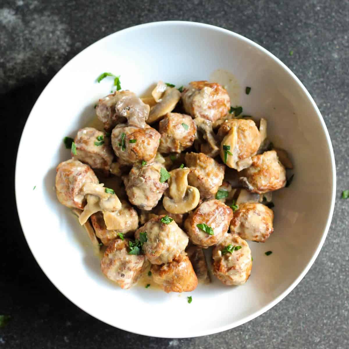 A white bowl filled with meatballs, mushrooms, and creamy sauce garnished with chopped herbs.