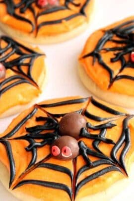 Cookies decorated with orange frosting, black spider web patterns, and chocolate spiders with red eyes. The cookies are arranged closely together.
