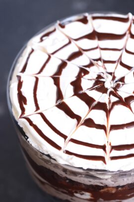 A close-up view of a dessert topped with whipped cream decorated with a spiderweb pattern made of chocolate syrup.