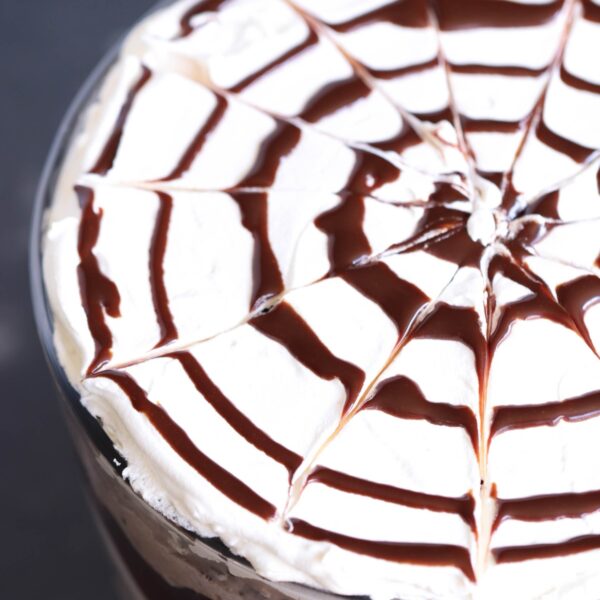 A close-up view of a dessert topped with whipped cream decorated with a spiderweb pattern made of chocolate syrup.