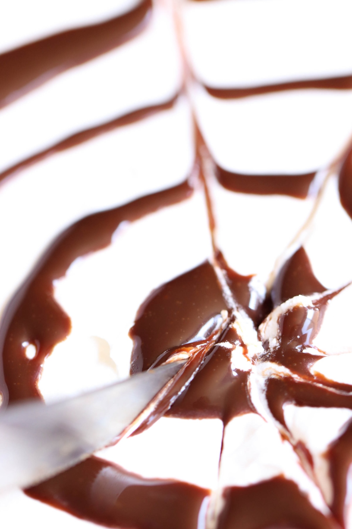 A close-up of a dessert with a knife cutting through a white surface drizzled with chocolate syrup in a star-like pattern.