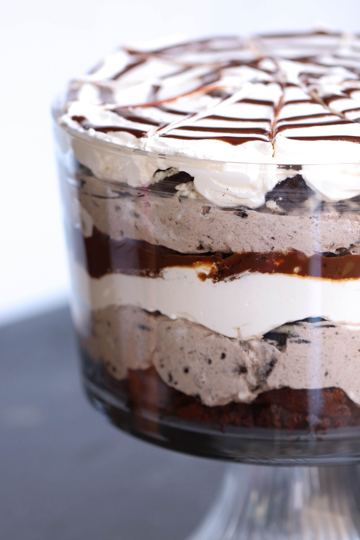 A layered trifle in a glass bowl, consisting of whipped cream, chocolate pudding, cake pieces, and a chocolate drizzle on top.