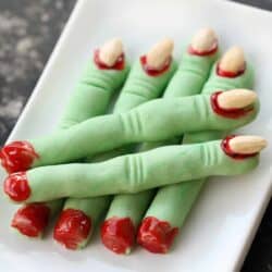 Plate of green finger-shaped cookies with almond nails and red icing detail, resembling severed fingers, on a white rectangular dish.