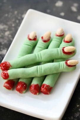 Plate of green finger-shaped cookies with almond nails and red icing detail, resembling severed fingers, on a white rectangular dish.