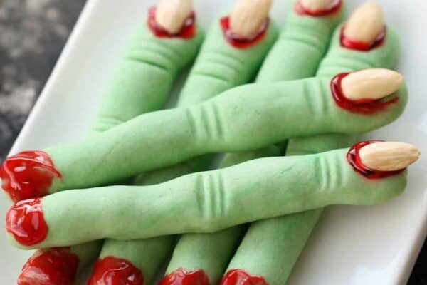 Plate of green finger-shaped cookies with almond nails and red icing detail, resembling severed fingers, on a white rectangular dish.