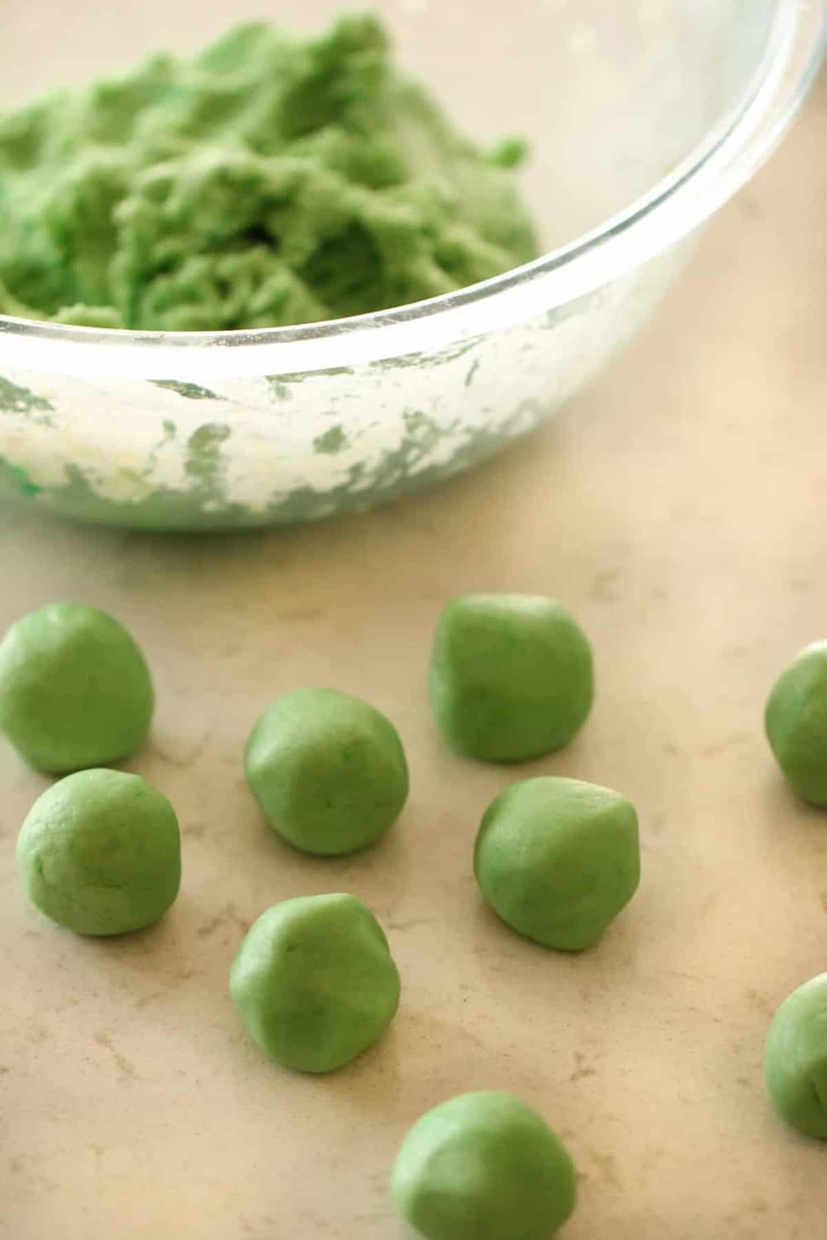 Close-up of a bowl of green dough in the background and several green dough balls placed on a countertop in the foreground.