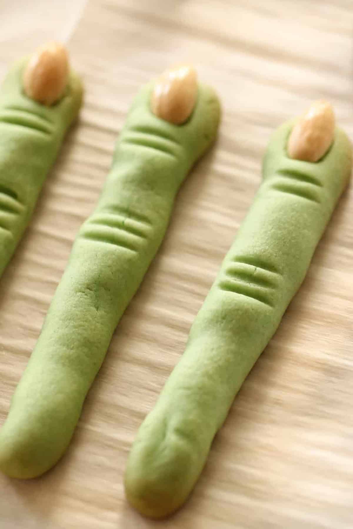 Three green, finger-shaped cookies with almond slivers to resemble fingernails, placed on a beige textured surface.