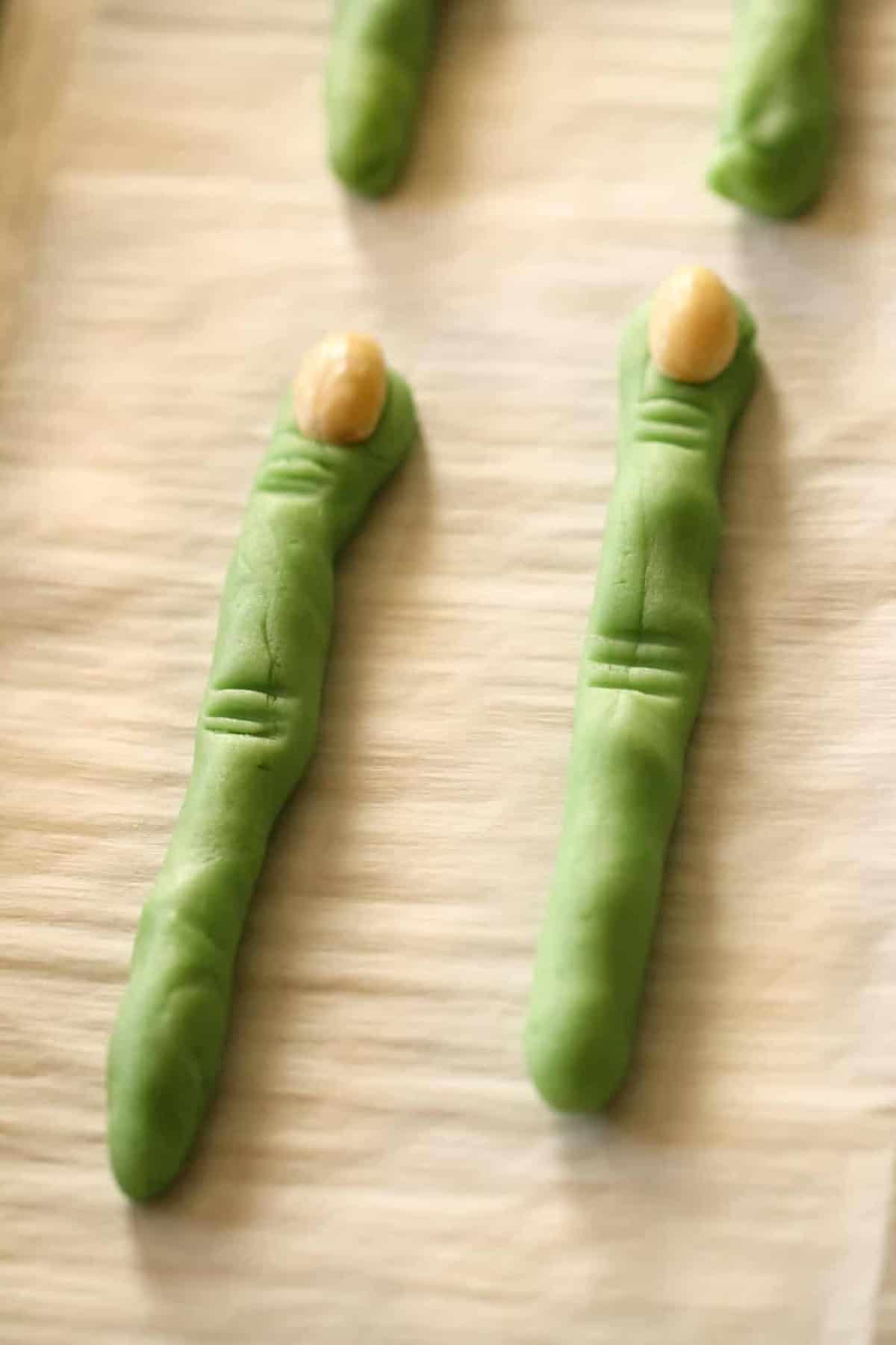 Two green cookie sticks shaped like fingers with almond halves as nails, placed on a parchment-lined baking sheet.