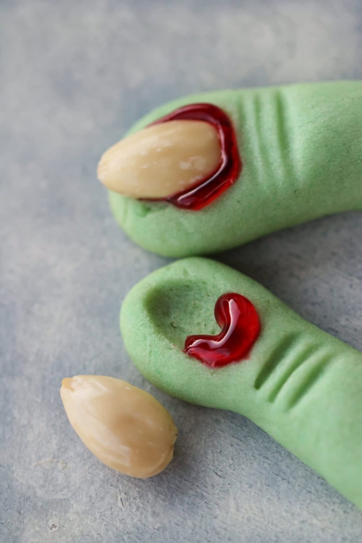 Close-up of green, finger-shaped cookies with a white almond resembling a fingernail; one almond is placed below the finger, with red icing mimicking blood.