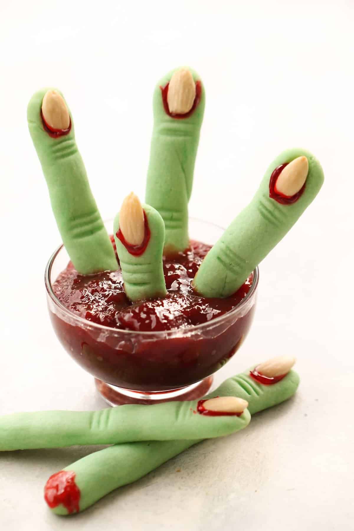 Green witch finger cookies with almond nails are dipped in red jam and arranged in a glass, resembling severed fingers. Additional cookies lie beside the glass on a light background.