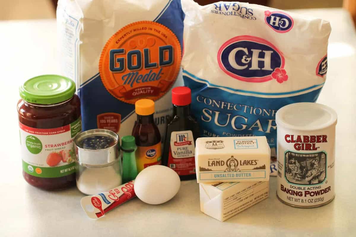 Assortment of baking ingredients on a counter, including flour, sugar, butter, baking powder, vanilla extract, food coloring, an egg, a jar of strawberry spread, salt, and a container of green sprinkles.