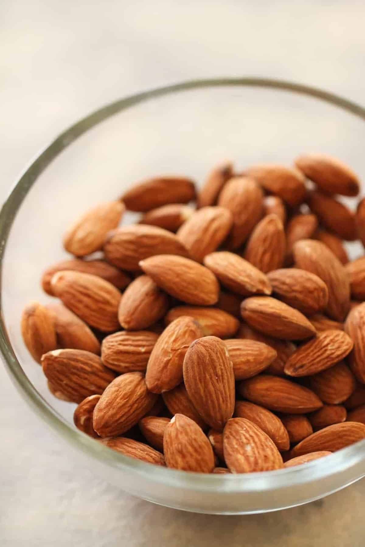 A glass bowl filled with whole, unshelled almonds placed on a light-colored surface.