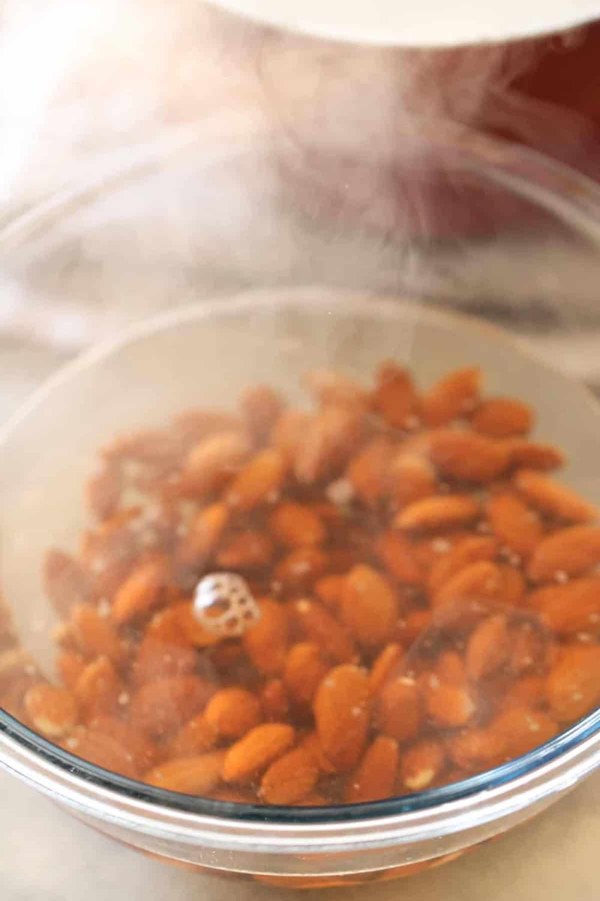 A glass bowl filled with almonds submerged in hot water, with steam rising from the surface.