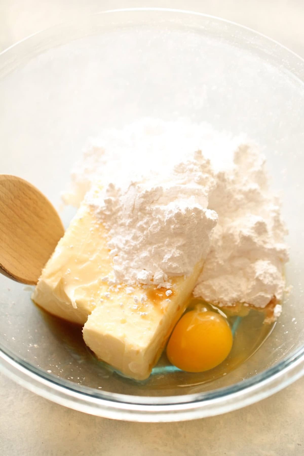 A clear mixing bowl containing two sticks of butter, a single egg, a wooden spoon, and a large quantity of powdered sugar.