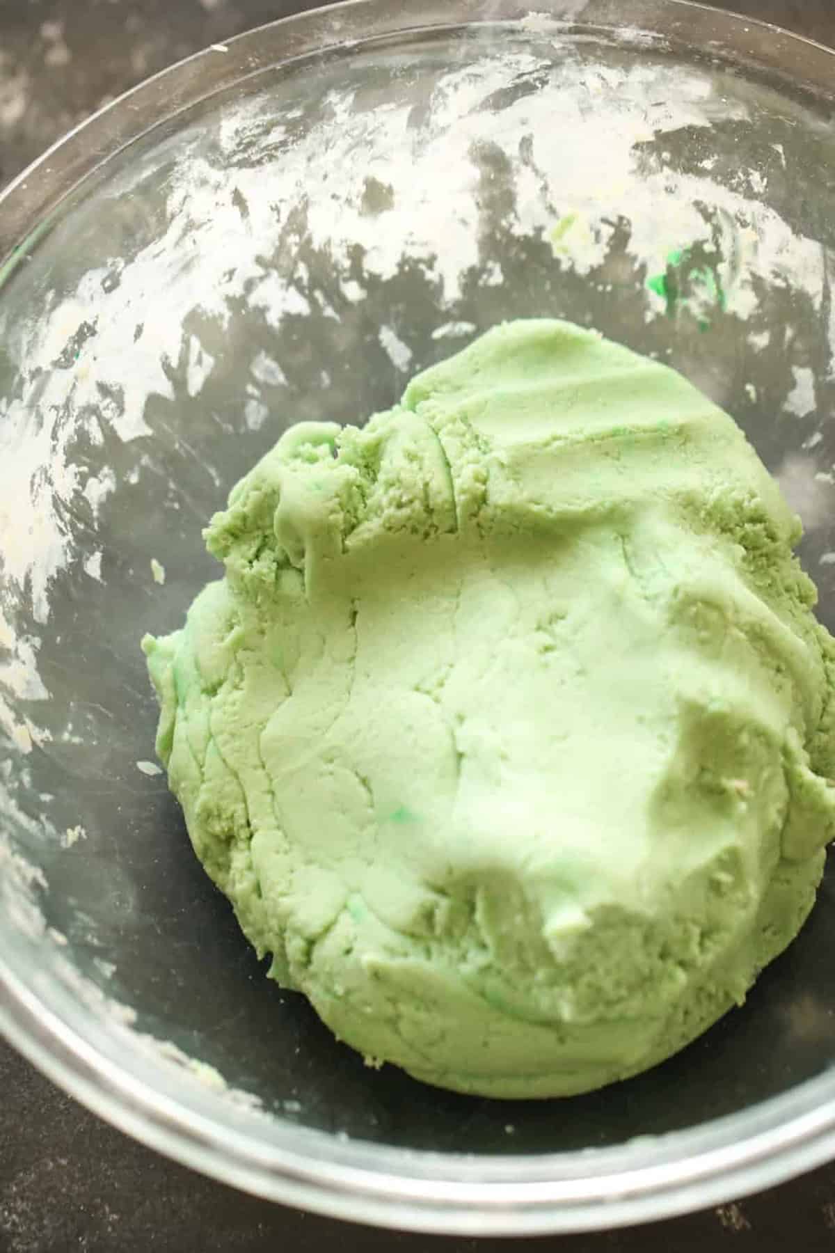 A clear glass bowl containing a mound of light green dough on a dark surface.