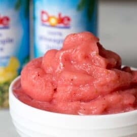 A white bowl holds pink strawberry Dole Whip, with two stacked cans of Dole crushed pineapple in the background.