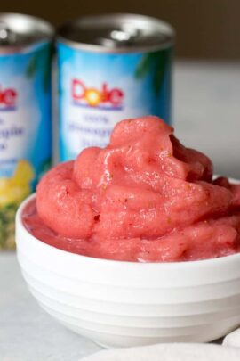 A white bowl holds pink strawberry Dole Whip, with two stacked cans of Dole crushed pineapple in the background.