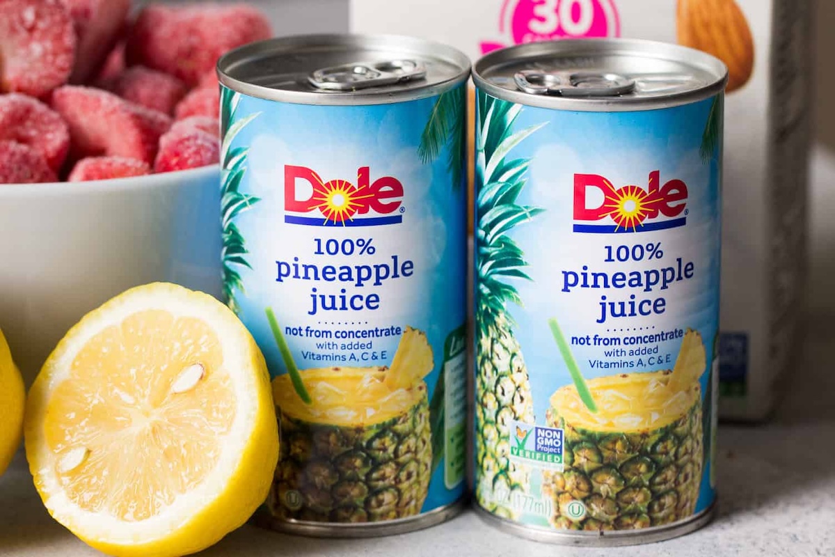 Two cans of Dole 100% pineapple juice are placed next to a halved lemon and a bowl of frozen strawberries.