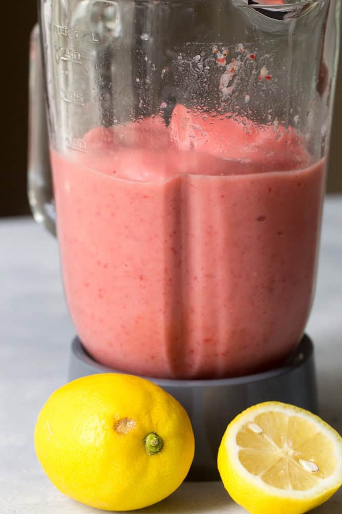 A blender containing pink strawberry smoothie mixture next to one whole lemon and one halved lemon on a surface.