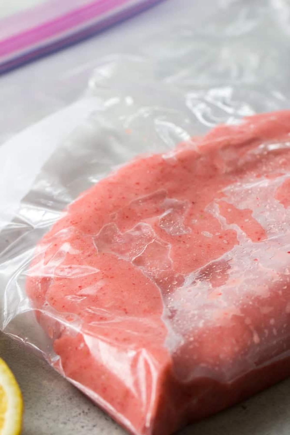 A close-up of a clear plastic bag filled with a pink, blended substance, possibly a fruit smoothie or puree, lying on a light-colored surface.
