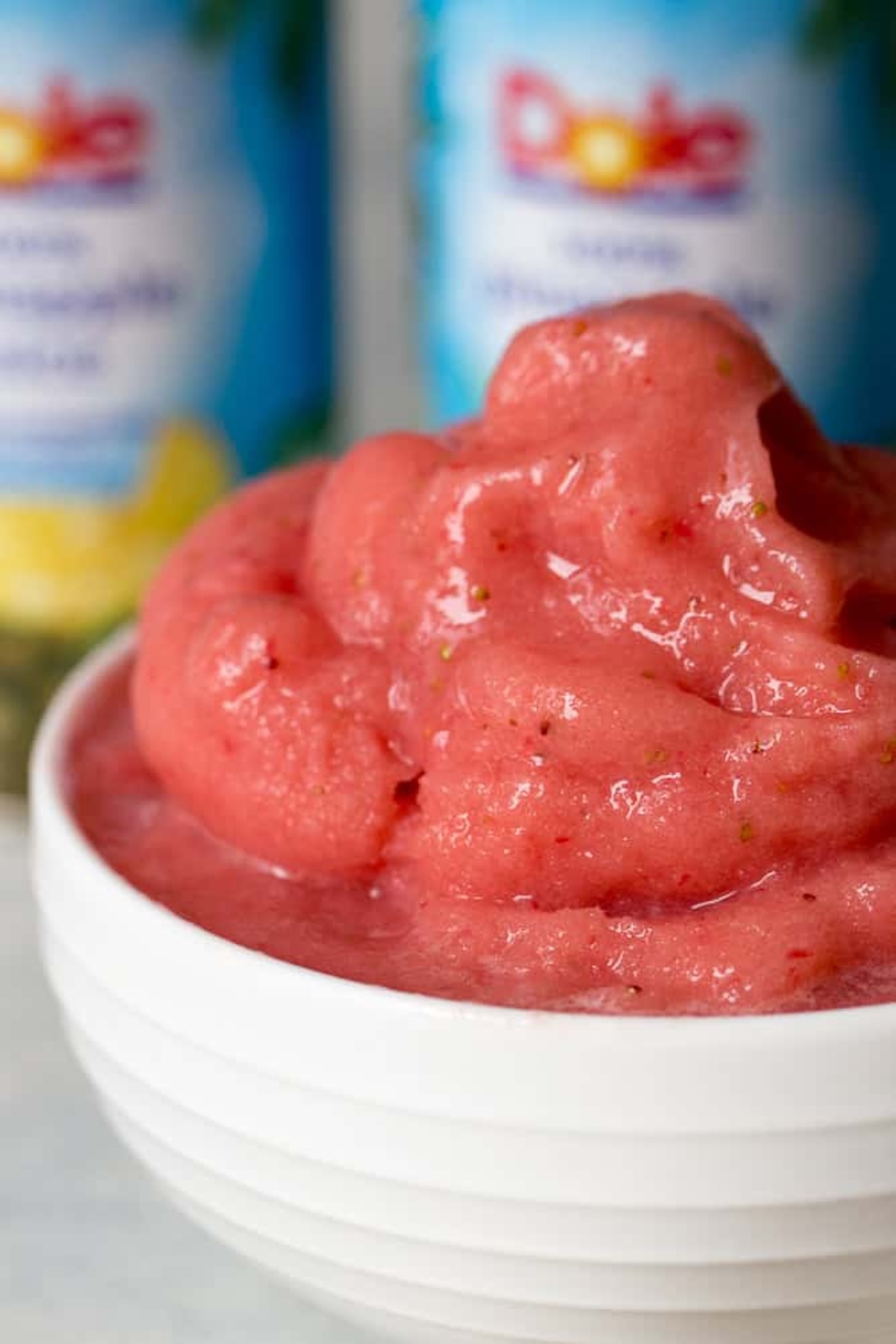 A white bowl filled with smooth, pink strawberry sorbet. In the blurred background, two cans of Dole pineapple juice are visible.