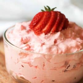 A glass bowl filled with pink strawberry mousse topped with a sliced strawberry, placed on a wooden surface.