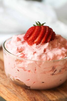 A glass bowl filled with pink strawberry mousse topped with a sliced strawberry, placed on a wooden surface.