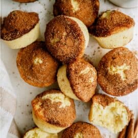 A pile of muffins with a cinnamon sugar topping is shown, some whole and some cut in half, displayed on a white surface with a cup of milk and cinnamon sticks nearby.