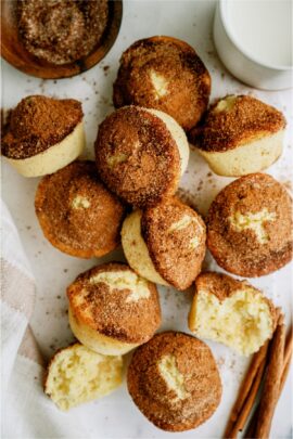 A pile of muffins with a cinnamon sugar topping is shown, some whole and some cut in half, displayed on a white surface with a cup of milk and cinnamon sticks nearby.