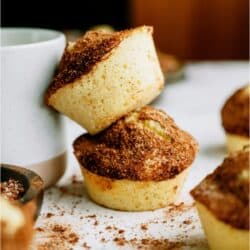 Two cinnamon sugar muffins are stacked beside a white cup, with more muffins and cinnamon sugar scattered on a white surface in the background.