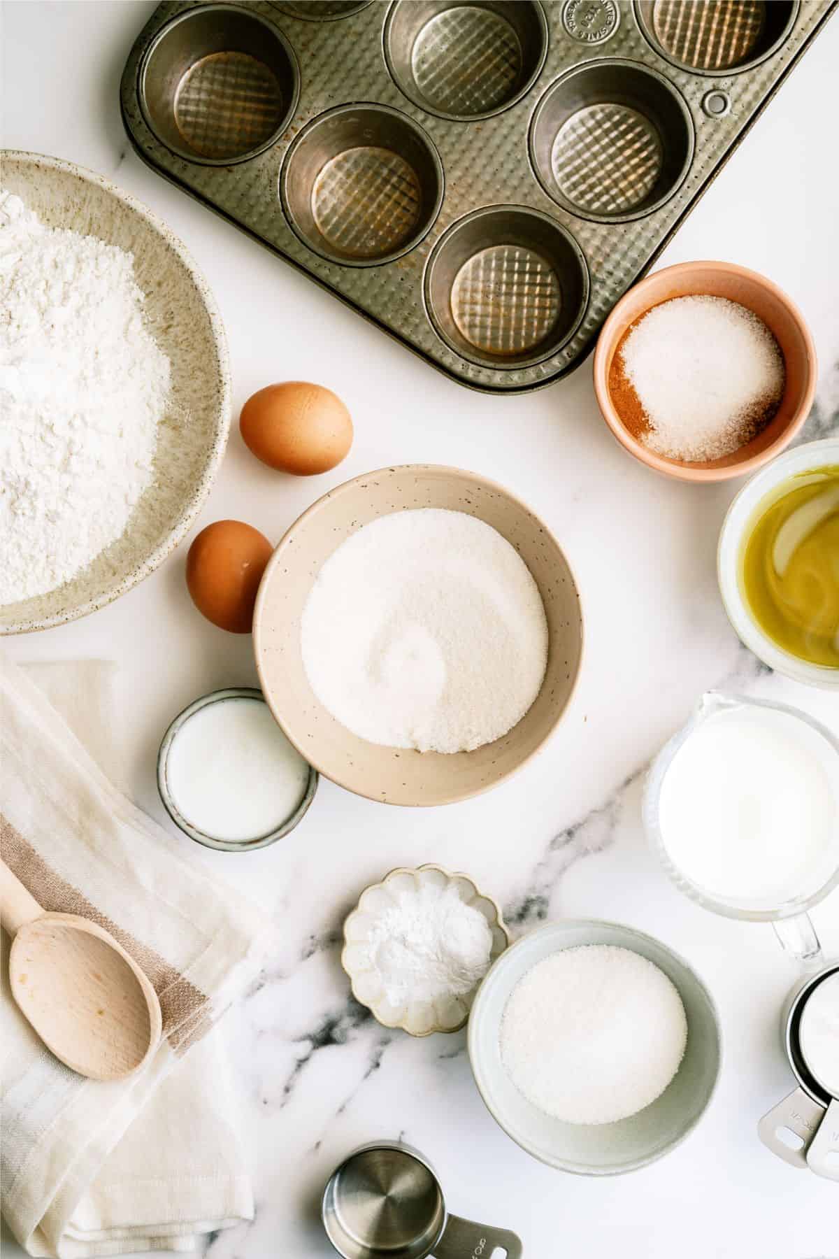 A top-down view of baking ingredients including flour, sugar, eggs, oil, milk, baking powder, and muffin tin, arranged on a white surface.