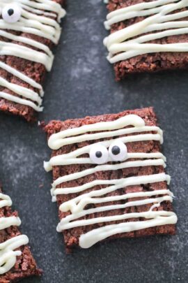 Brownies topped with white icing, resembling mummies, with candy eyes on a dark surface.