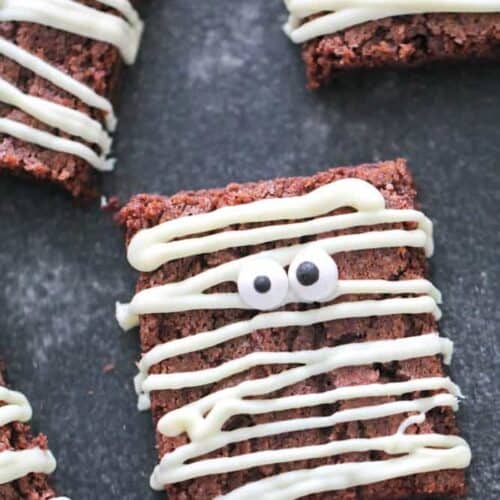 Brownies topped with white icing, resembling mummies, with candy eyes on a dark surface.