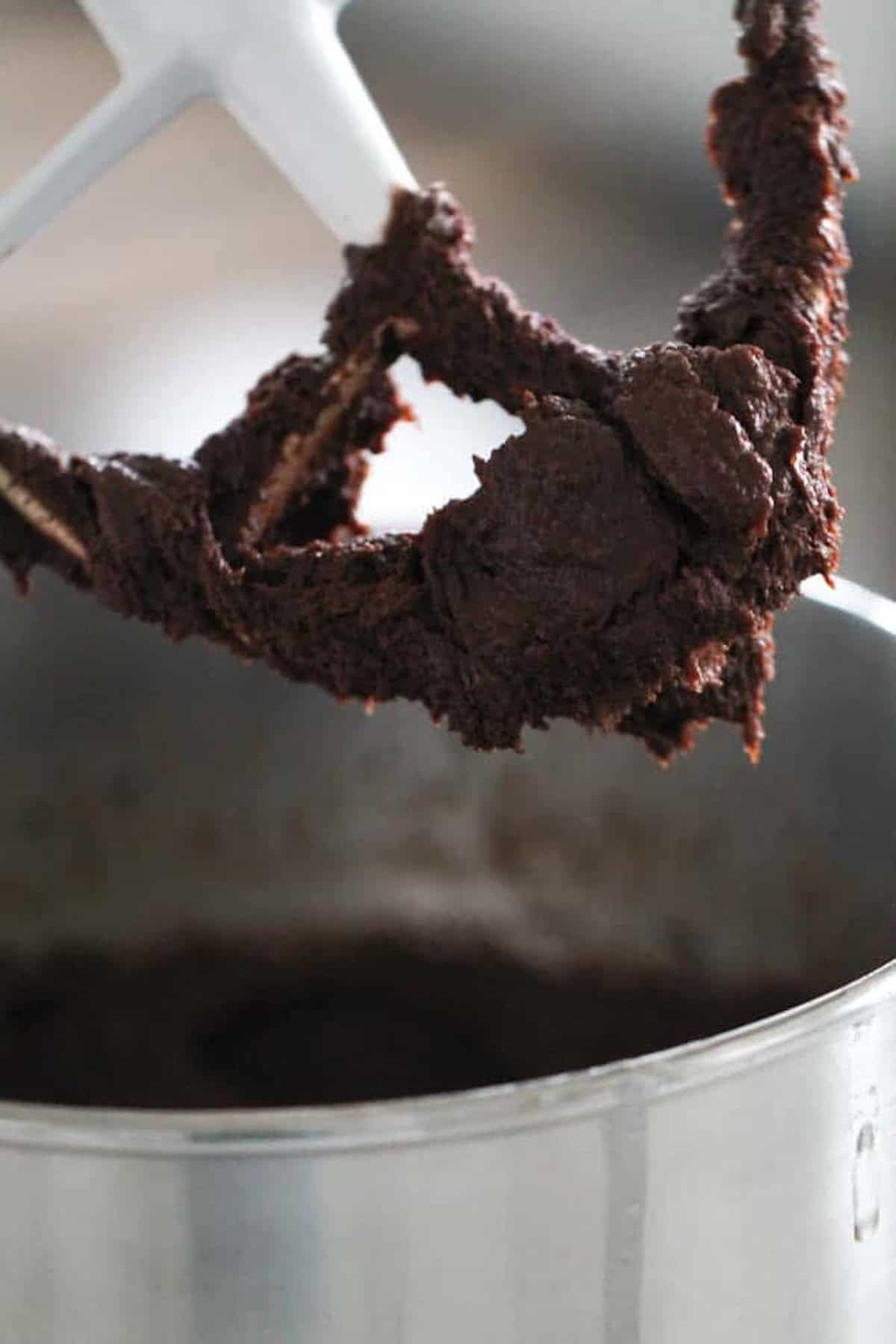 Close-up of a stand mixer's paddle attachment coated in thick chocolate batter, with a metal mixing bowl partially visible below.