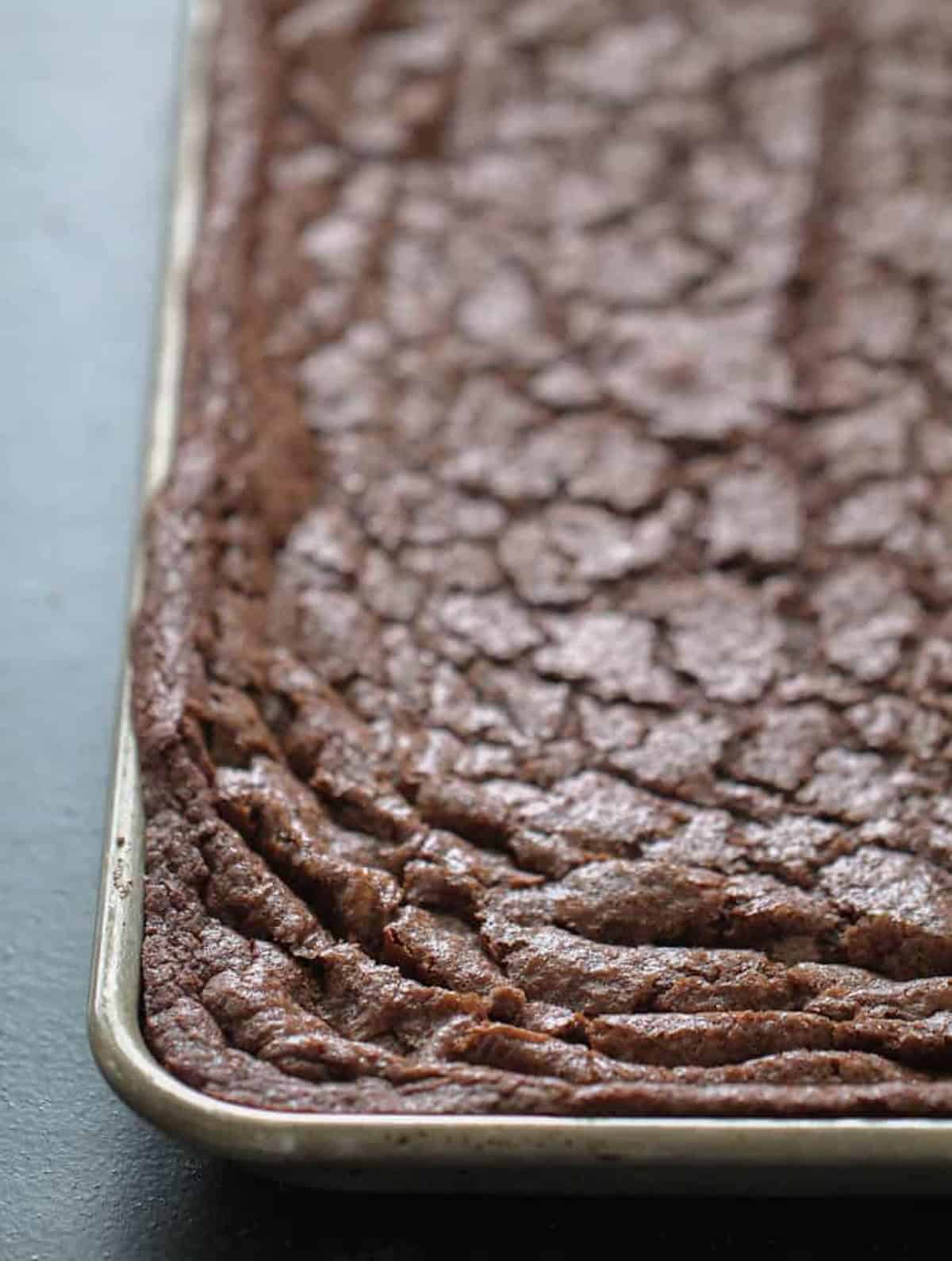 A close-up of a large rectangular baking tray filled with freshly baked brownies, showing a cracked, crispy surface.