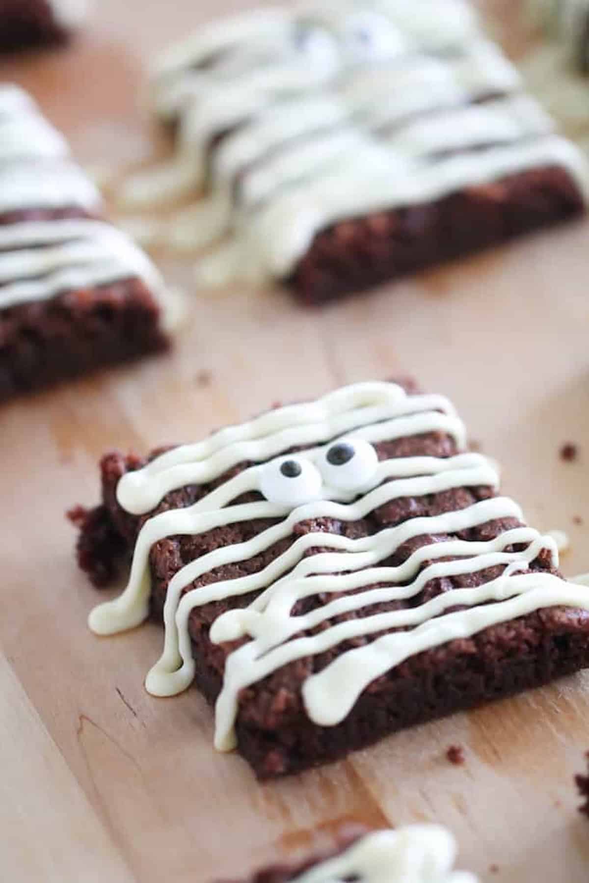 Brownies with white icing drizzled in such a way that it resembles a mummy, complete with candy eyes on a wooden surface.