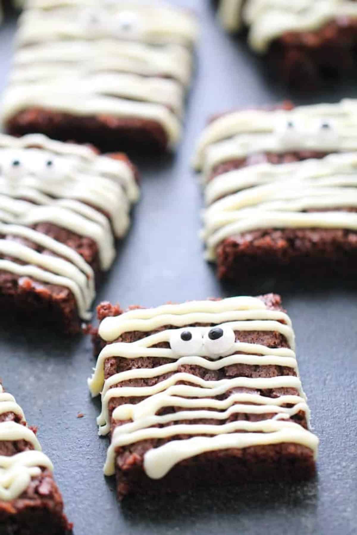 Brownie squares decorated with white icing to resemble mummies, with two candy eyes on each, placed on a dark surface.