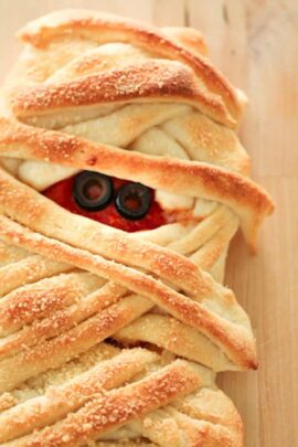 A baked pastry shaped like a mummy with two sliced black olives representing eyes peeking out from between the pastry layers.