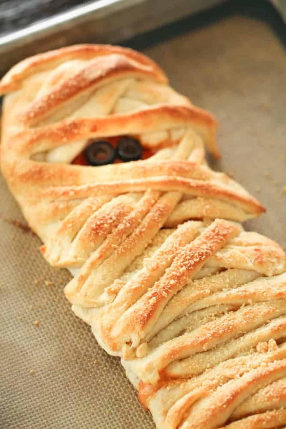 A loaf of bread is baked to resemble a mummy, with olive slices used as eyes, placed on a baking sheet.